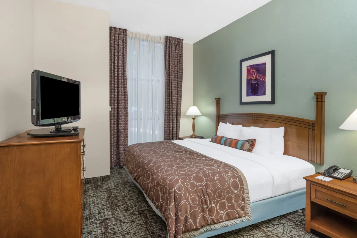 A bedroom scene featuring a neatly made bed with decorative pillows, a television set positioned for easy viewing, and a cozy nightstand with a lamp. The soft lighting and inviting textures create a comforting retreat perfect for relaxation or enjoying a movie night.