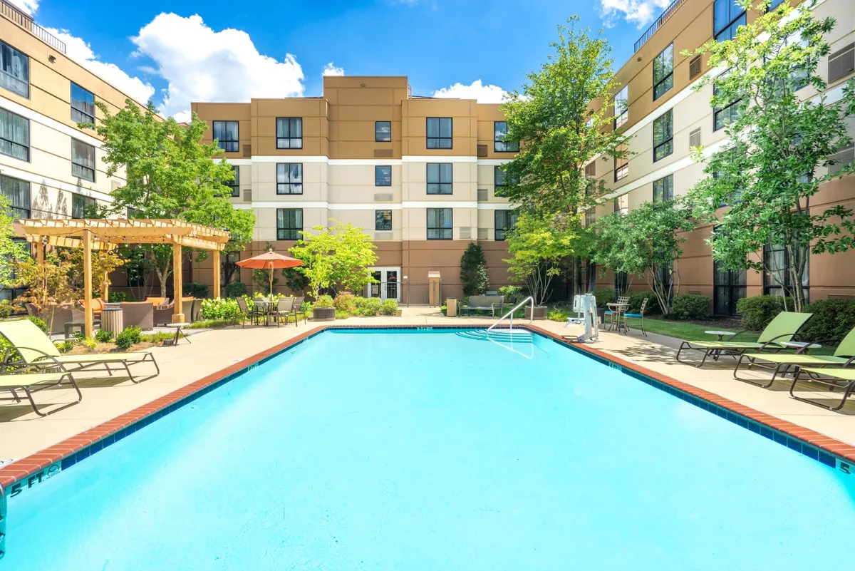 An inviting exterior pool area surrounded by lounge chairs, a pergola with seating, and lush green trees. Guests are seen swimming, sunbathing, and socializing under the pergola, all set against the backdrop of a building facade. The sunset lighting adds a cheerful, relaxed mood.