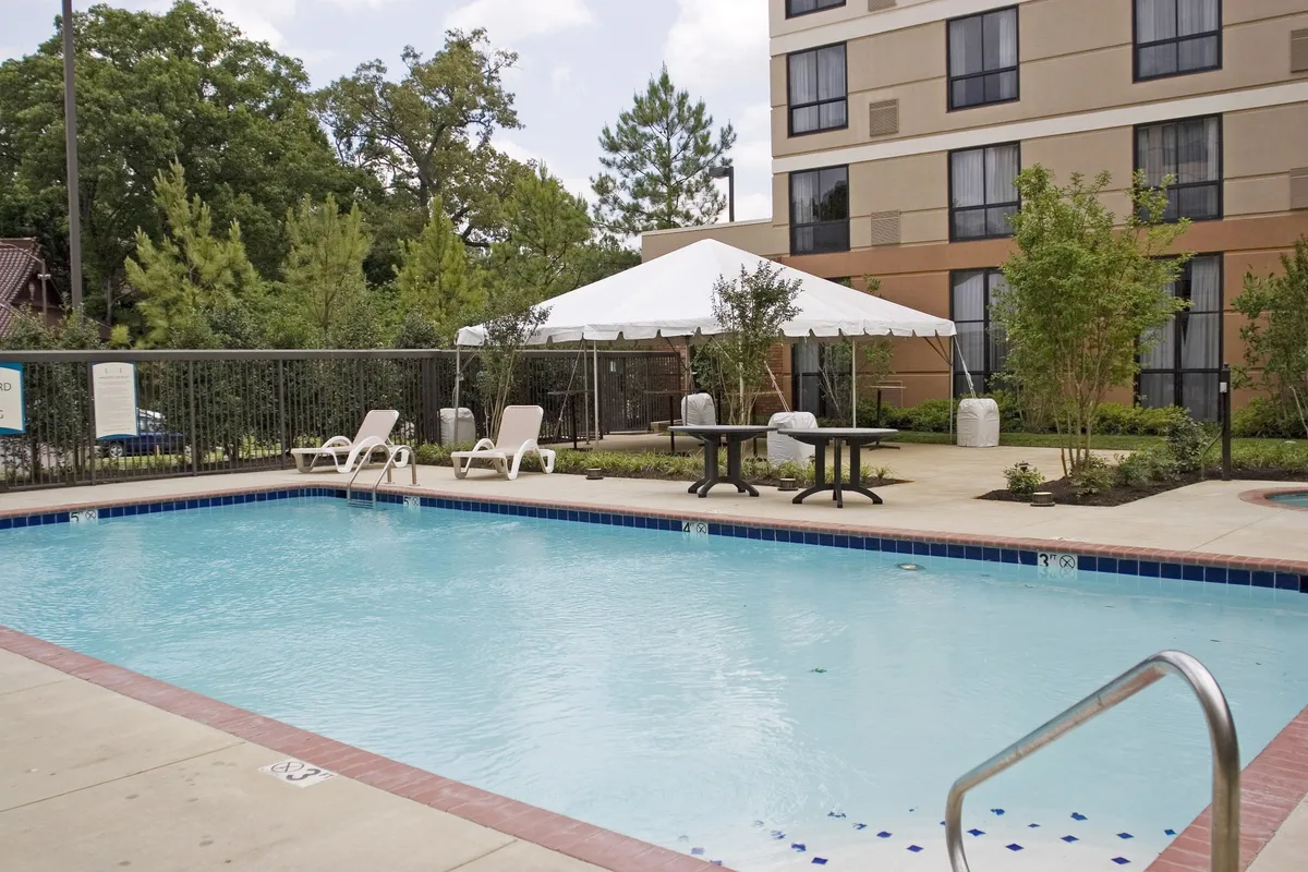 A bright outdoor pool area with clear blue water, lounge chairs lining the deck, a tented seating area, and lush landscaping creating a welcoming environment for relaxation and fun. The sunny weather and inviting setup hint at opportunities for family activities and downtime after a day of exploring local attractions.