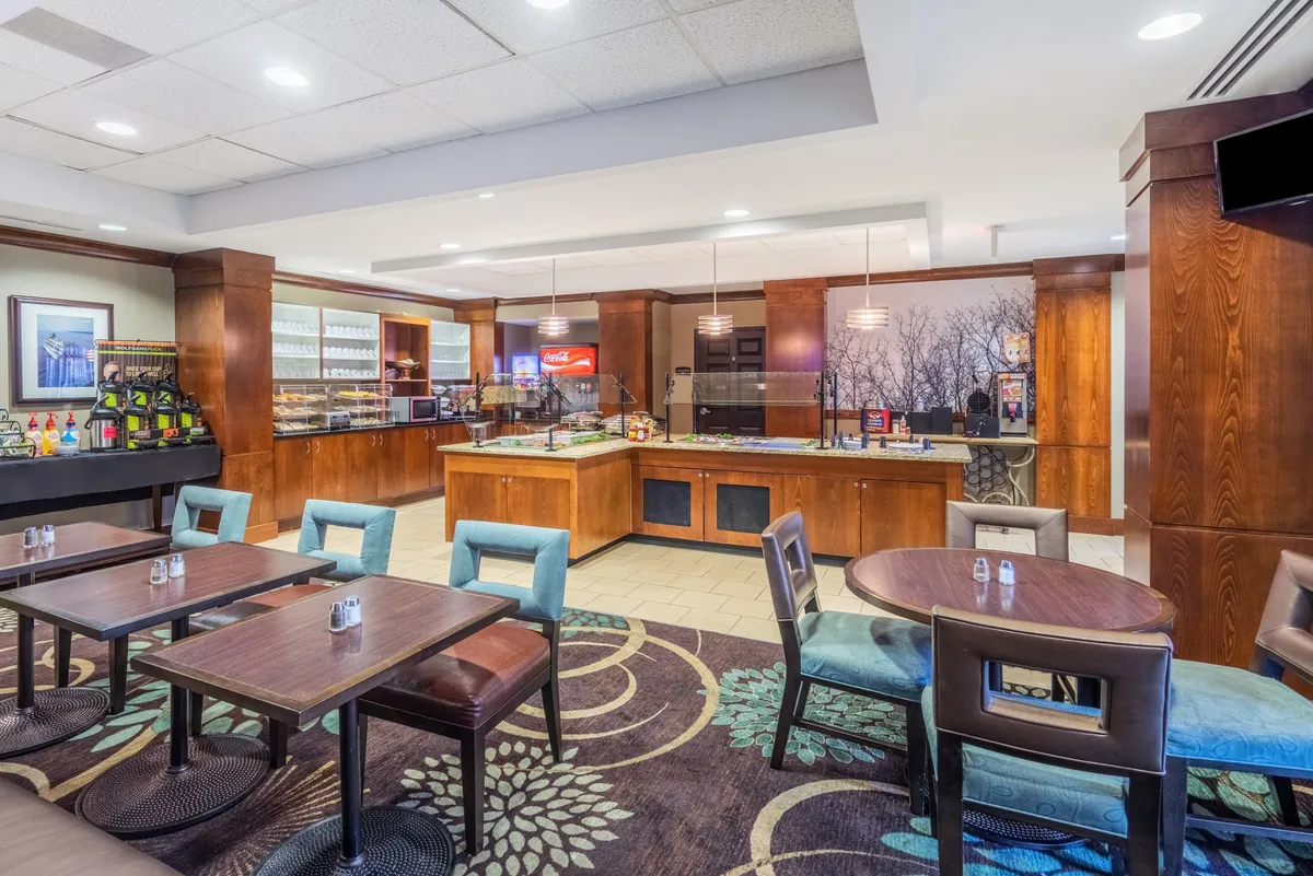This image shows a contemporary wooden counter with a seating area, tasteful wall art, and a beverage station. The space is designed for casual dining or working, offering a functional yet comfortable environment suitable for productivity and socializing.
