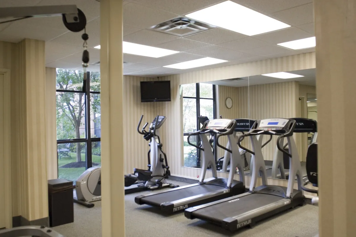 A bright, modern fitness center featuring several treadmills and an elliptical machine, set against large windows that fill the space with natural light. The equipment is arranged for both individual workouts and group exercise, with a TV screen for entertainment and ample space for users to move comfortably.