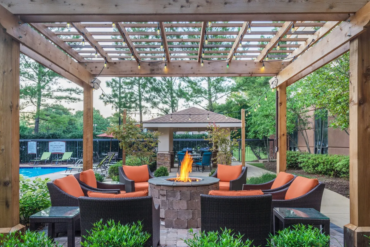 An inviting exterior patio space featuring a cozy fire pit surrounded by a comfortable seating arrangement and a wooden pergola. The area is set up for outdoor social gatherings and barbecues, capturing a warm, communal atmosphere ideal for sharing meals.